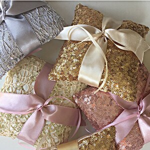 May include: Four ring bearer pillows with different designs. One pillow is silver with a lace design and a white satin ribbon. Another pillow is gold with a lace design and a pink satin ribbon. The third pillow is gold with sequins and a cream satin ribbon. The fourth pillow is rose gold with sequins and a pink satin ribbon.
