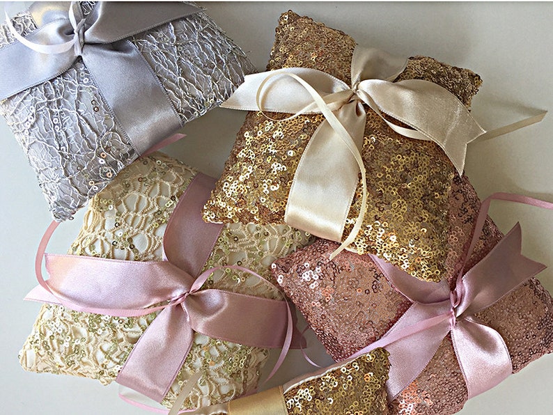 May include: Four ring bearer pillows with different designs. One pillow is silver with a lace design and a white satin ribbon. Another pillow is gold with a lace design and a pink satin ribbon. The third pillow is gold with sequins and a cream satin ribbon. The fourth pillow is rose gold with sequins and a pink satin ribbon.