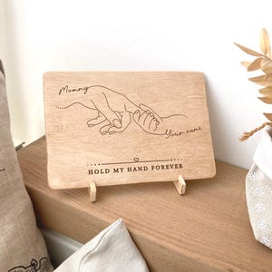 May include: Wooden sign with a hand-drawn illustration of two hands holding each other. The text on the sign reads "Mommy" and "Your name" and "Hold my hand forever".