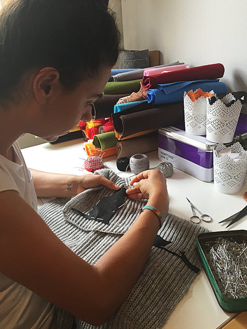 Puede incluir: Una persona est&aacute; cosiendo un parche de tela negro en un su&eacute;ter de punto gris. Hay rollos de fieltro de colores, carretes de hilo y una lata de alfileres de coser en la mesa.