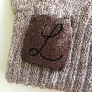 Puede incluir: Un gorro de punto marr&oacute;n con un parche de cuero marr&oacute;n que tiene la letra "L" bordada en hilo negro.