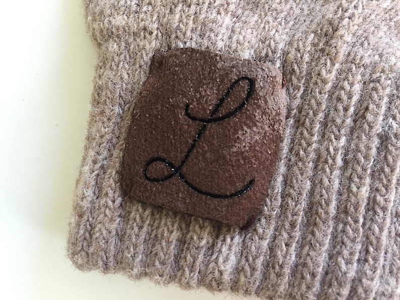 Puede incluir: Un gorro de punto marr&oacute;n con un parche de cuero marr&oacute;n que tiene la letra "L" bordada en hilo negro.