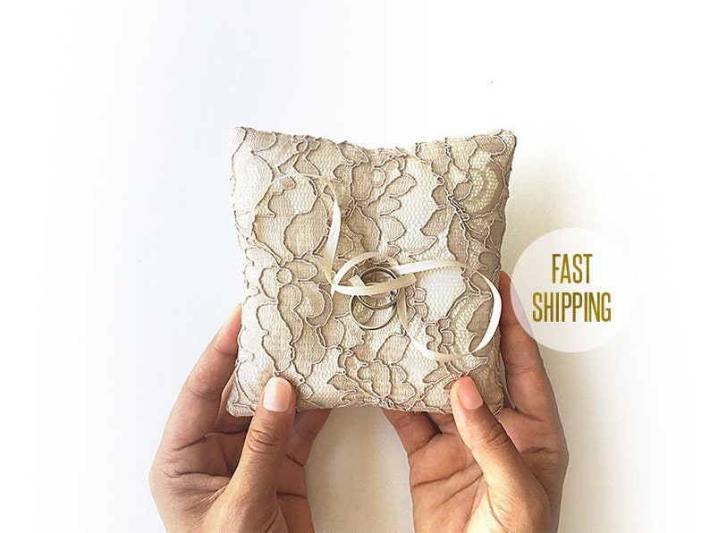 May include: A white lace ring pillow with two gold rings tied with a white ribbon. The pillow is being held by two hands.