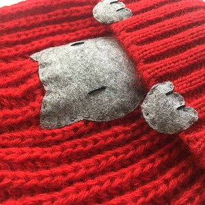 Puede incluir: Sombrero de punto rojo con orejas de gato de fieltro gris y una cara de gato de fieltro gris en la parte delantera.