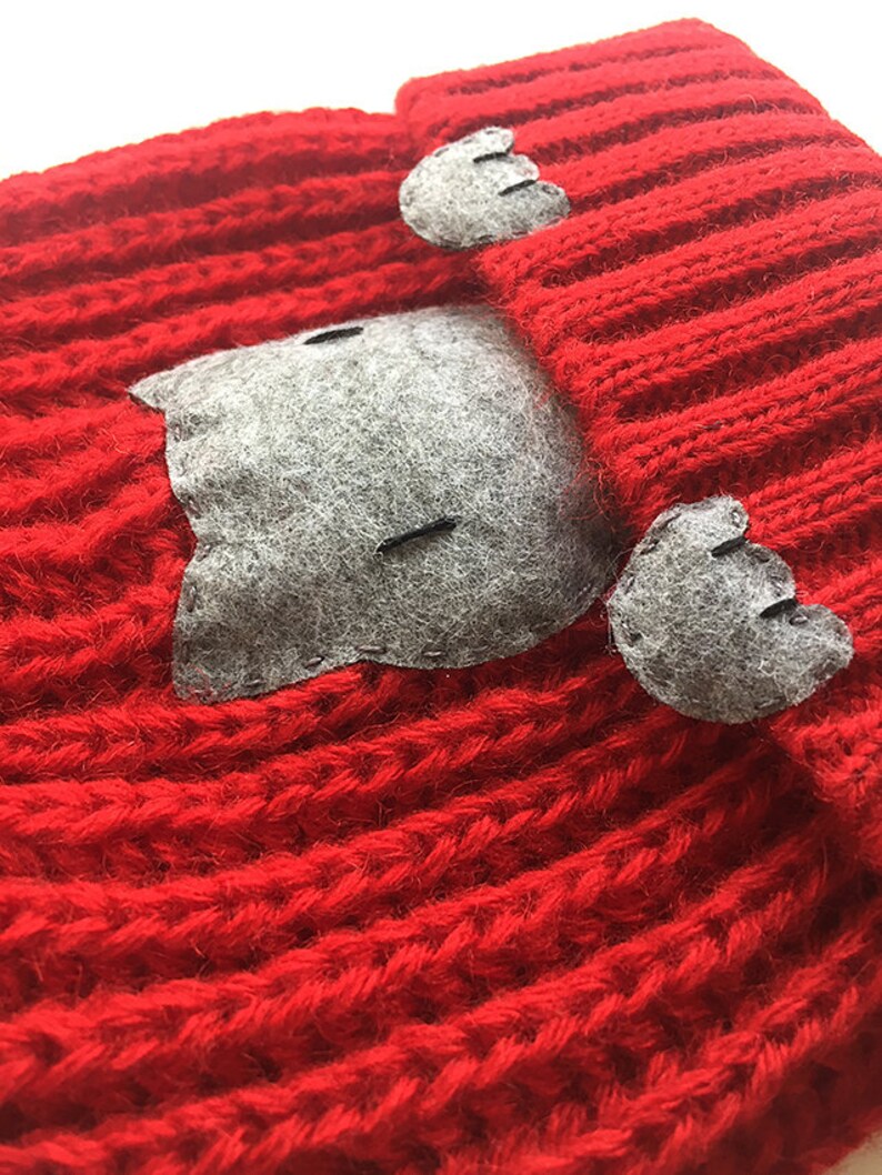 Puede incluir: Sombrero de punto rojo con orejas de gato de fieltro gris y una cara de gato de fieltro gris en la parte delantera.