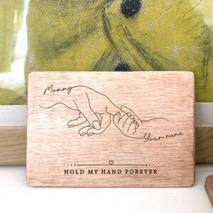 May include: A wooden plaque with a hand-drawn illustration of two hands holding each other. The text "Mommy" and "Your name" are engraved on the plaque, along with the phrase "Hold my hand forever".