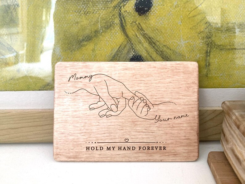May include: A wooden plaque with a hand-drawn illustration of two hands holding each other. The text "Mommy" and "Your name" are engraved on the plaque, along with the phrase "Hold my hand forever".