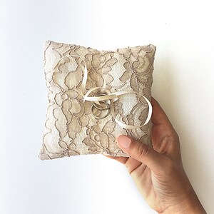 May include: A small, white lace pillow with a satin ribbon tied around it. Two gold rings are placed on the pillow.