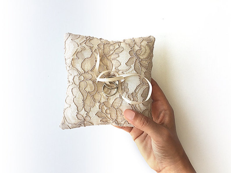 May include: A small, white lace pillow with a satin ribbon tied around it. Two gold rings are placed on the pillow.