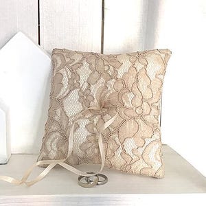 May include: A beige lace ring pillow with a satin ribbon and two gold wedding bands. The pillow is sitting on a white shelf.