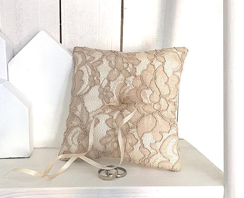 May include: A beige lace ring pillow with a satin ribbon and two gold wedding bands. The pillow is sitting on a white shelf.