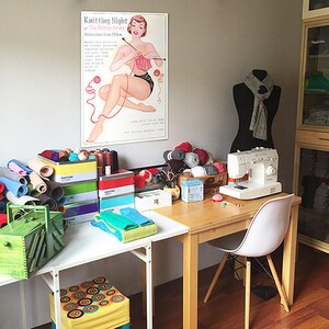 Puede incluir: Una sala de costura con una mesa blanca, una m&aacute;quina de coser, un maniqu&iacute; y un cartel que dice "Knitting Night". Tambi&eacute;n hay varias bolas de hilo de colores, rollos de tela y otros materiales de costura.