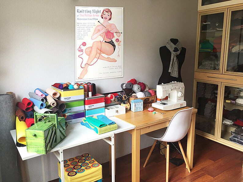 Puede incluir: Una sala de costura con una mesa blanca, una m&aacute;quina de coser, un maniqu&iacute; y un cartel que dice "Knitting Night". Tambi&eacute;n hay varias bolas de hilo de colores, rollos de tela y otros materiales de costura.