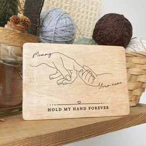 May include: A wooden sign with a line drawing of two hands holding each other. The text on the sign reads "Mommy" and "Your name" with the words "Hold my hand forever" below the drawing.