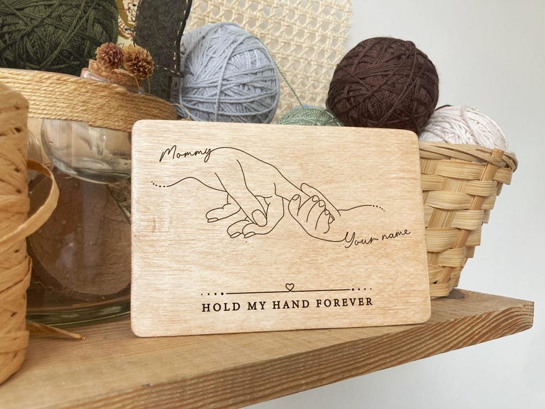 May include: A wooden sign with a line drawing of two hands holding each other. The text on the sign reads "Mommy" and "Your name" with the words "Hold my hand forever" below the drawing.