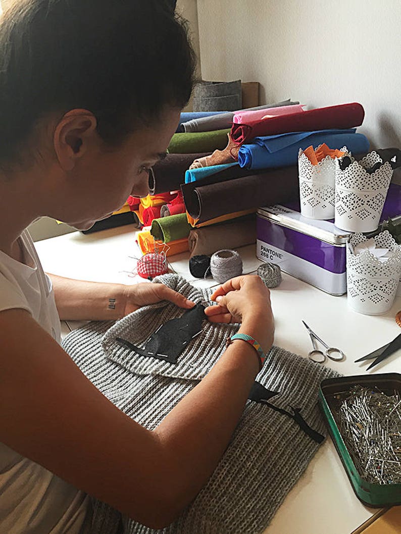 Puede incluir: Una persona est&aacute; cosiendo un parche negro en un su&eacute;ter de punto gris. Hay varios colores de tela de fieltro enrollados en el fondo, junto con una caja de alfileres de coser y un carrete de hilo.