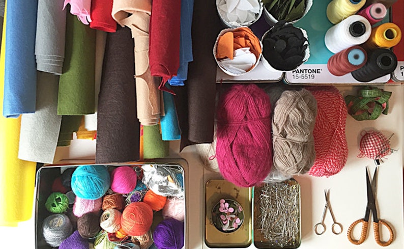 Puede incluir: Una colecci&oacute;n de hilo, fieltro e hilo de colores, incluyendo rosa, rojo, azul, verde y marr&oacute;n. Los materiales est&aacute;n organizados en contenedores y sobre una superficie blanca. La imagen tambi&eacute;n incluye materiales de costura, como tijeras y alfileres.