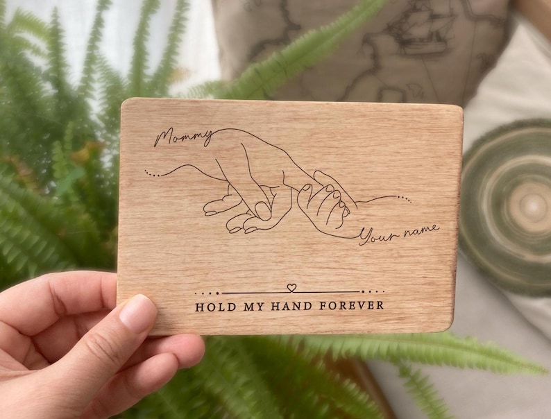 May include: Wooden plaque with a hand-drawn illustration of two hands holding each other. The text "Mommy" and "Your name" are engraved on the plaque. The text "Hold my hand forever" is also engraved on the plaque.