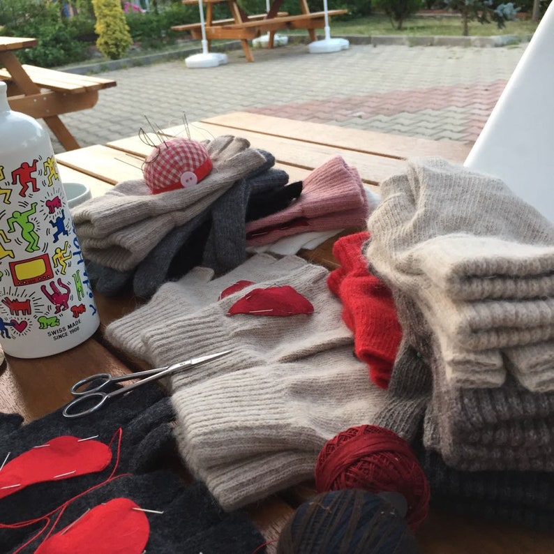 Puede incluir: Una mesa de madera con una variedad de art&iacute;culos de punto, incluyendo guantes, bufandas y sombreros. Hay un trozo de tela rojo en forma de coraz&oacute;n en uno de los guantes y una bola de lana roja en la mesa. Tambi&eacute;n hay un par de tijeras y un alfiletero con un patr&oacute;n de cuadros rojos y blancos.