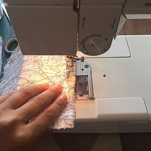 May include: A white sewing machine with a hand holding a piece of white fabric with a silver pattern. The fabric is being fed into the machine.