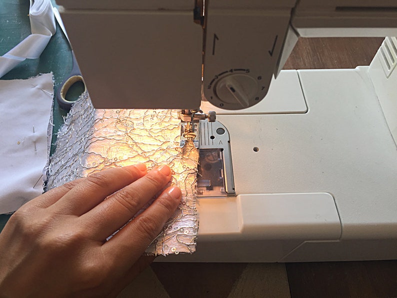 May include: A white sewing machine with a hand holding a piece of white fabric with a silver pattern. The fabric is being fed into the machine.