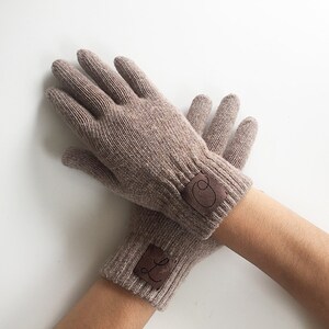 Puede incluir: Un par de guantes de punto marr&oacute;n con un parche de cuero marr&oacute;n en cada guante. Los parches tienen las letras "L" y "O" en ellos.