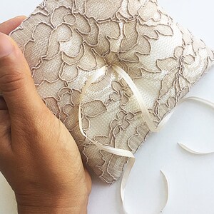May include: A beige lace ring pillow with a satin ribbon tied in a bow. The pillow is held in a hand.