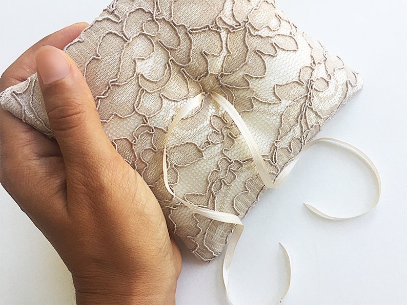 May include: A beige lace ring pillow with a satin ribbon tied in a bow. The pillow is held in a hand.