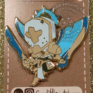 May include: Enamel pin of a cartoon knight with a white helmet and blue details, wielding two swords. The pin is displayed on a brown card with the text "ENAMEL PINS" and social media details. The background is a gold glitter surface.