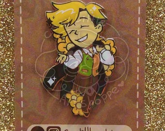 Op de afbeelding: Een emaille pin met een cartoonfiguur met blond haar, een groen vest en een donkere cape, bevestigd op een bruine kaart met de tekst ENAMEL PINS by cozylittleartshoppe, inclusief sociale media iconen en de tekst Cozylittleartshoppe en Cozylittleartblog.