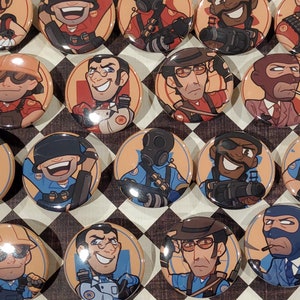 May include: A set of 16 round button pins featuring cartoon characters from the video game Team Fortress 2. The pins are on a checkered black and white surface.