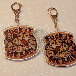 May include: Two clear acrylic keychains with gold-colored hardware. Each keychain features a cartoon snake design in shades of brown and tan, with the text "FUCK IT WE BALL" in brown lettering. The keychains are on a light tan surface.