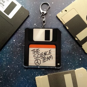 May include: A black and orange floppy disk keychain with the text "The Science Team" and a number 1 on it. The keychain is attached to a silver key ring.