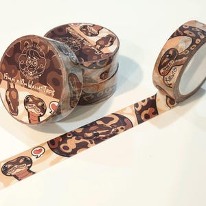 Op de afbeelding: Verschillende rollen washi tape met een slangenpatroon en de tekst "Cozy Little Artshoppe". De tape is 15 mm x 10 m. E&eacute;n rol is afgerold en toont het slangenontwerp. De tape heeft een bruin, beige en cr&egrave;me kleurenschema.