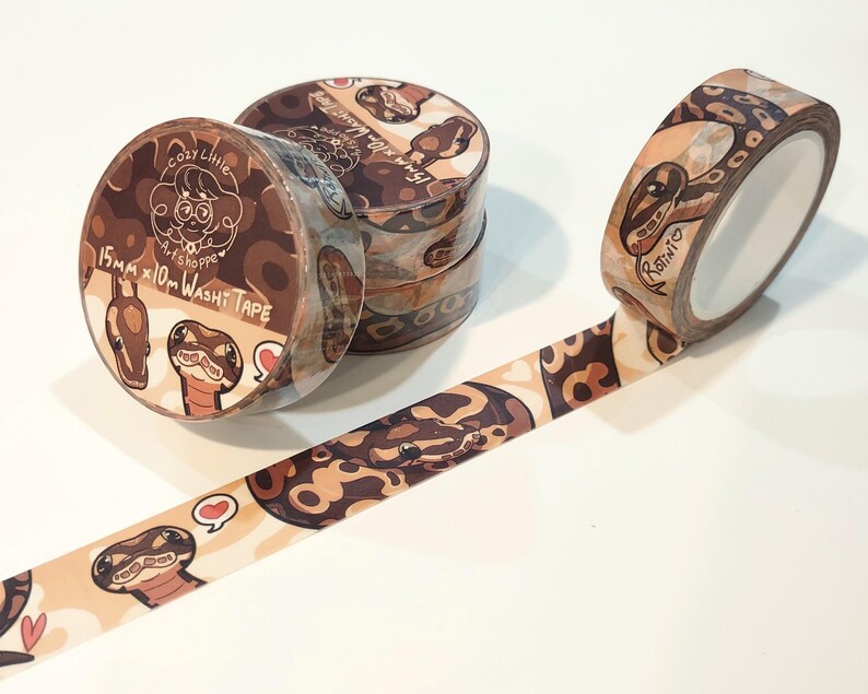 Op de afbeelding: Verschillende rollen washi tape met een slangenpatroon en de tekst "Cozy Little Artshoppe". De tape is 15 mm x 10 m. E&eacute;n rol is afgerold en toont het slangenontwerp. De tape heeft een bruin, beige en cr&egrave;me kleurenschema.