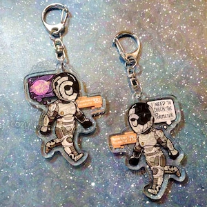 May include: Two clear acrylic keychains featuring a cartoon astronaut design. One keychain has a thought bubble with the text "I NEED TO CHECK THE PERIMETER." The other has a thought bubble with a planet graphic and the text "Supernova Thursday."