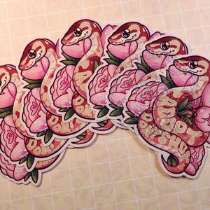 May include: A collection of die-cut stickers featuring a tan and red snake coiled around pink peony flowers. The snake has large black eyes and the text "ADOPT DON'T SHOP" is integrated into its body pattern in a light cream color.