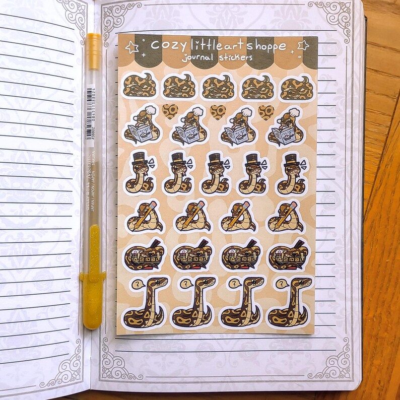 Op de afbeelding: Een vel met journalstickers met cartoon slangen in verschillende poses, waaronder lezen, het dragen van hoge hoeden en het vasthouden van potloden. De stickers staan op een bruinachtige achtergrond met de tekst "cozy little art shoppe journal stickers". Een gouden pen ligt op een open notitieboek.