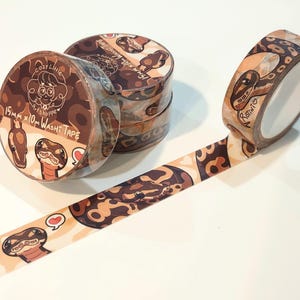 May include: Washi tape rolls with a snake pattern in shades of brown and tan. The tape is 15mm x 10m and features illustrations of snakes and the text "Cozy Little Art Shoppe". One roll is unrolled, showing the snake design.