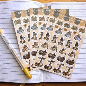 May include: Three sticker sheets with snake illustrations in various poses, including reading and wearing hats. The stickers are on a light brown background with the text "Cozy littleheart shoppe journal stickers." A gold pen rests on an open notebook.
