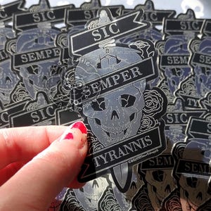 May include: A collection of stickers featuring a skull design with a dagger and banner. The banner reads "Semper Tyrannis" and "Sic". The stickers are primarily silver and black, with floral accents.