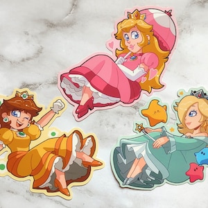 May include: Three cartoon character stickers featuring Princess Peach, Daisy, and Rosalina. Peach is wearing a pink dress and a mushroom hat. Daisy is wearing a yellow dress and a flower crown. Rosalina is wearing a blue dress and a crown. All three characters are smiling.