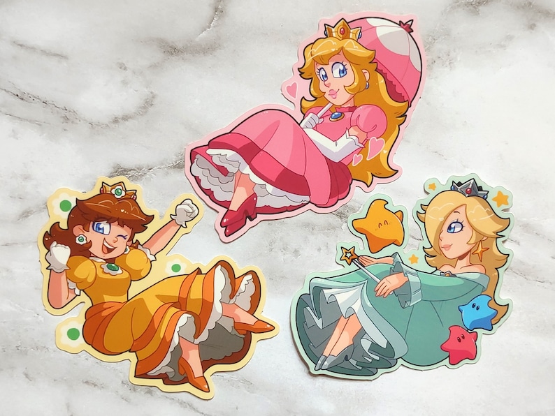 May include: Three cartoon character stickers featuring Princess Peach, Daisy, and Rosalina. Peach is wearing a pink dress and a mushroom hat. Daisy is wearing a yellow dress and a flower crown. Rosalina is wearing a blue dress and a crown. All three characters are smiling.