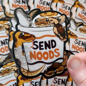 以下が含まれることがあります： 「SEND NOODS」の文字が入ったカップの中に、漫画のヘビが描かれたステッカー。ヘビは茶色、タン、白。ステッカーは手で持たれています。