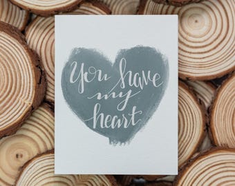 Puede incluir: Una tarjeta blanca con un corazón gris azulado pintado a mano. Las palabras "You have my heart" están escritas en escritura blanca dentro del corazón. La tarjeta está sobre un fondo de rodajas de madera.