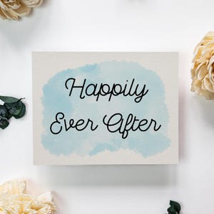 May include: A rectangular card with the words "Happily Ever After" in black script over a light blue watercolor wash. The card is surrounded by white flowers and greenery on a white background.