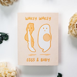Puede incluir: Una tarjeta de felicitación con un fondo melocotón claro presenta las palabras "WAKEY WAKEY" en naranja. Debajo del texto hay ilustraciones de un huevo y tocino. Las palabras "EGGS & BABY" están impresas en naranja debajo de las ilustraciones.
