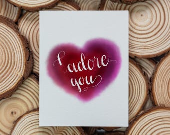 Puede incluir: Una tarjeta de felicitación rectangular blanca con una forma de corazón de color magenta y rojo oscuro con bordes suaves en el centro. El texto "I adore you" está escrito en blanco con una elegante letra cursiva.