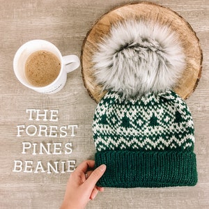 May include: A green and white knit beanie with a white faux fur pom pom. The beanie has a green band at the bottom. The beanie is on a wooden surface with the words "THE FOREST PINES BEANIE" written in white letters.