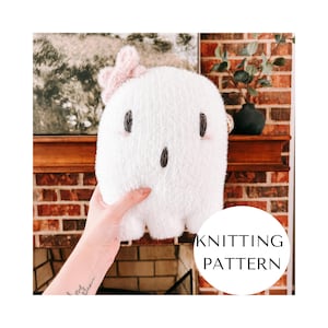 May include: A white plush ghost with a pink bow, black embroidered eyes and mouth, and pink cheeks. The ghost is a knitting pattern. The image is set against a brick wall and a wooden mantelpiece.